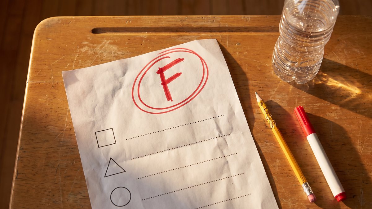 Illustration: a school quiz marked with a big red circled F on a desk, beside a plastic water bottle, a chewed pencil and a red marker