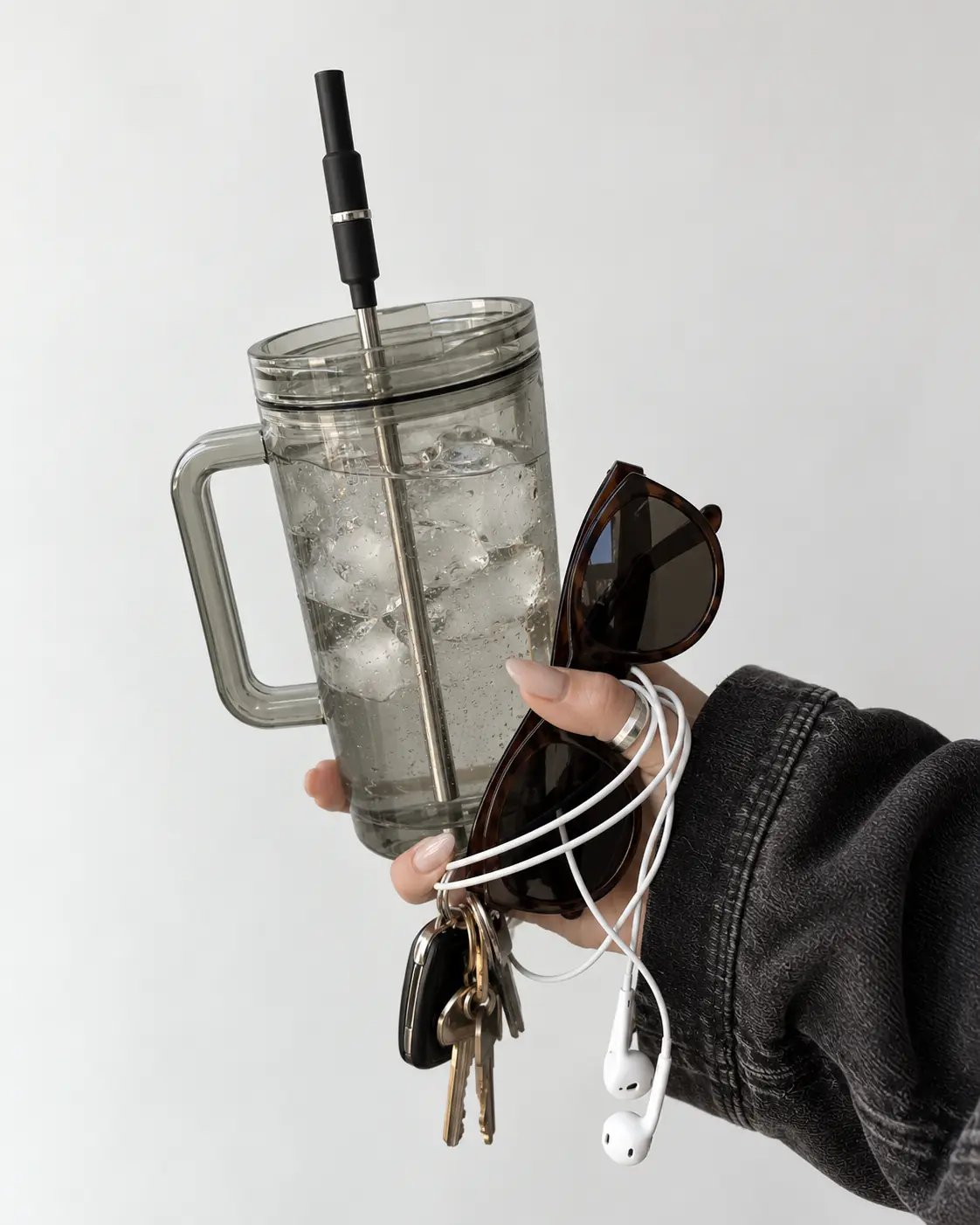 A hand holding a clear tumbler with a black Aelo Straw, sunglasses, keys and earphones.